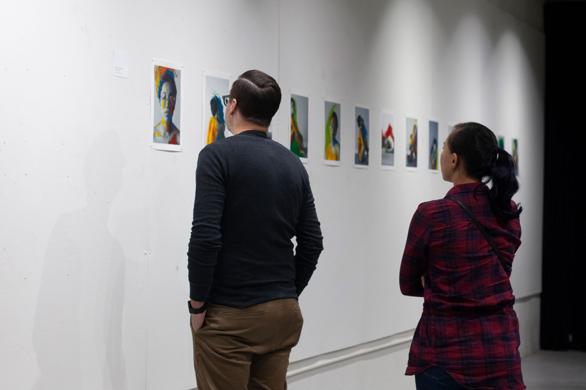 a gallery wall full of colorful paintings, two people looking at the series